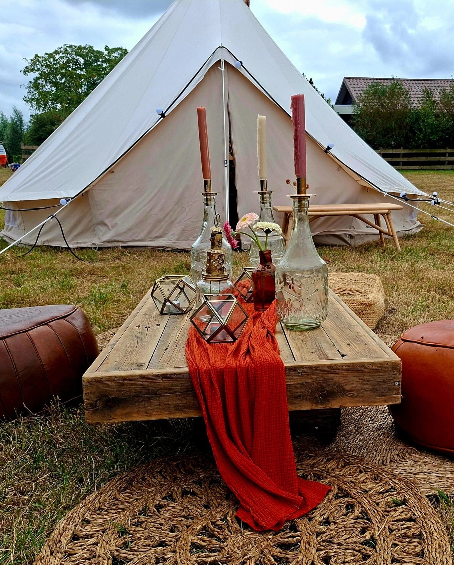 Lage houten tafel voor de tenten met kaarsen, glazen flessen, een oranje loper en leren poefs: het stylingpakket